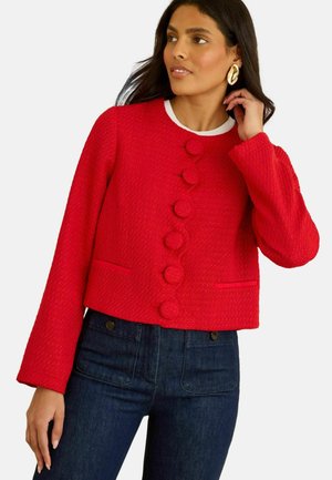 Red cropped jacket with textured fabric, featuring round buttons, long sleeves, and two front pockets, paired with dark jeans.