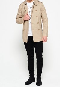 Cappotto beige con bottoni doppi, spalline e tasche laterali, abbinato a pantaloni slim neri e stivaletti alla caviglia.