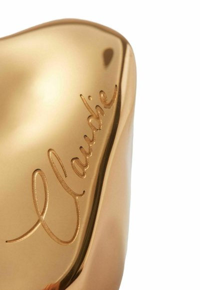 Objeto metálico de color dorado, suave al tacto y de forma redondeada, con texto en relieve que dice "Claudie" en un lado. La superficie reflectante resalta los detalles.