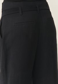 Gros plan sur un pantalon noir sur mesure avec poches passepoilées à l'arrière et passants pour ceinture sur un fond clair.