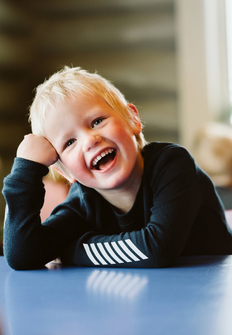 Bambino con capelli biondi corti, che indossa una maglietta nera a maniche lunghe con accenti a righe bianche, sdraiato su un tavolo blu, mentre sorride ampiamente.