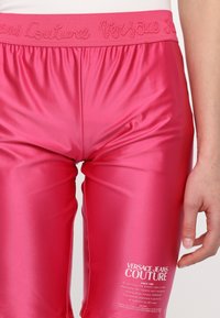Růžové saténové tepláky s elastickým pasem zdobené výšivkou "Versace Couture". Logo a textový detail na dolní části nohy.
