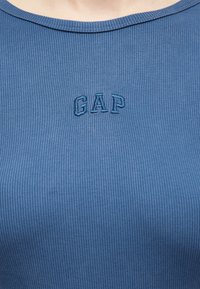 Κοντινό πλάνο ενός ατόμου που φοράει μπλε τοπ από ριμπ ύφασμα με το κέντημα "GAP" στο στήθος με μπλε κλωστή.