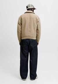 Chaqueta bomber de mezcla de lana color beige con puños y cuello acanalados, combinada con unos pantalones de mezclilla de pierna ancha azul oscuro. La vista trasera no muestra detalles visibles.