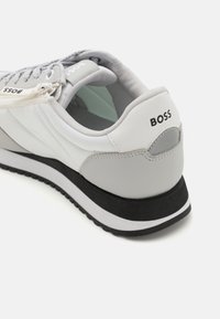 Valkoinen ja harmaa tennari, jossa on sekoitus tekstiiliä ja nahkaa. Siinä on harmaa kantapää, musta ulkopohja ja "BOSS"-logo takana.