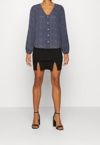 Blusa blu con pois bianchi, apertura frontale con bottoni e maniche a palloncino abbinata a una gonna nera aderente con spacchi laterali. Sandali neri con tacco.