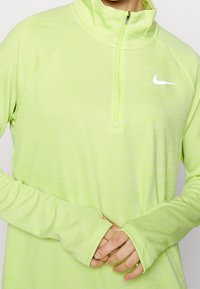 Jasnozielony polar Nike, zapinany na suwak do połowy, o miękkiej fakturze, z długimi rękawami z otworami na kciuki oraz odblaskowym logo na piersi.