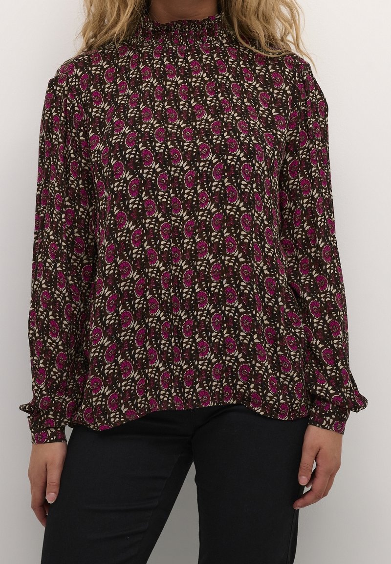 Blouse marron foncé à manches longues avec un col froncé, ornée d'un motif floral rose et crème et d'une texture de tissu douce. Un pantalon noir est porté en dessous.