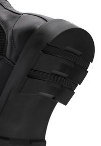 Bota de cuero negro con un acabado texturizado, que presenta una suela de goma gruesa con patrones de tracción estriados y un tacón redondeado.