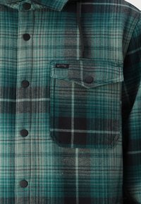 Flanellskjorta i teal, svart och ljusgrå rutmönster; har svarta knappar, en bröstficka och en stiliserad etikett.