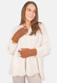 Junge Frau, die braune gestrickte Handschuhe und eine weiße grob gestrickte Strickjacke trägt, lächelt und schaut leicht zur Seite.