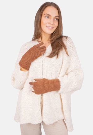 Young woman wearing brown knitted gloves and a white chunky knit cardigan, smiling and looking slightly to the side.