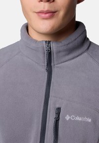 Grau fleecejacke mit hohem Kragen, Frontreißverschluss und dezentem Logo. Strukturierter Stoff und ein Reißverschluss-Taschendetail.