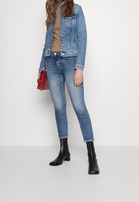 Chaqueta de mezclilla con un silueta ajustada, lavado azul claro, cierre frontal con botones, combinada con jeans azul de cintura alta y botines negros. Incluye un bolso rojo.