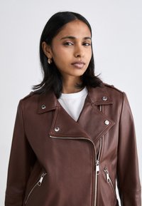 Jachetă de motociclist din piele maro, cu elemente metalice argintii, buzunare cu fermoar și rever. Textură netedă și croială ajustată.