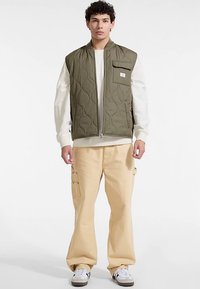 Olívazöld steppelt mellény elülső cipzárral és mellzsebbel, világos bézs cargo nadrággal és fehér tornacipővel párosítva. Simább anyagtextúra.