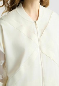 Lehká krémová bunda se zipem vpředu, vybavená průhlednými panely a diagonálním prošíváním pro moderní design.