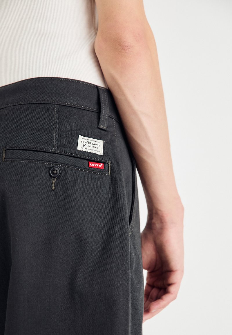 Pantalones gris oscuro hechos de una tela de textura suave. Presentan un bolsillo trasero con una etiqueta blanca y acentos rojos. Diseño sencillo.