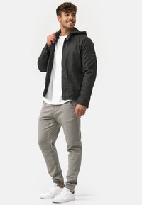 Schwarze Lederjacke mit Kapuze, texturierten Schultern, kombiniert mit hellgrauen, schmal zulaufenden Hosen und weißen Sneakers, vor einem schlichten Hintergrund stehend.