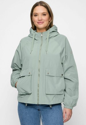 Vrouw draagt een lichtgroene capuchonjas met een ritssluiting aan de voorkant en twee klepzakken, gecombineerd met een blauwe spijkerbroek, staand tegen een witte achtergrond.