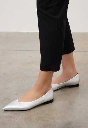 Füße, die spitze silberne flache Schuhe tragen, kombiniert mit schwarzen gekürzten Hosen, stehend auf einem Betonboden.