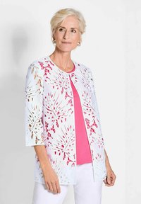 Cardigan en dentelle blanc avec des découpes en forme de feuilles, sur un top rose. Il présente des manches 3/4 et une coupe droite, offrant un design léger et aéré.