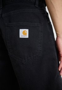 Sorte denim bukser med en lomme og en hvid label, der viser et gult logo, pænt syet detaljer og en glat tekstur.