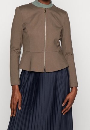Femme portant une veste zippée taupe avec une légère taille peplum sur une jupe plissée bleu marine, les mains relaxées sur les côtés.