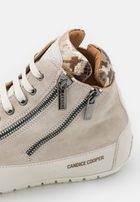 Béžové semišové vysoké tenisky s bílými tkaničkami, dvojicí černých zipů označených "CANDICE" a límcem se vzorem hada v zlaté a hnědé barvě.