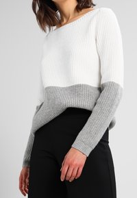 Sweter dziany z białą i szarą kolorystyką blokową, charakteryzujący się prążkowaną fakturą i długimi rękawami. Noszony z czarnymi spodniami.