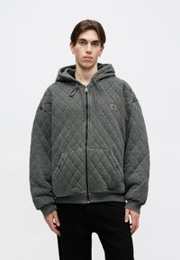 Sudadera con capucha acolchada de tela gris oscuro, que cuenta con un zipper completo, bolsillos frontales y un pequeño parche con logo en el pecho.