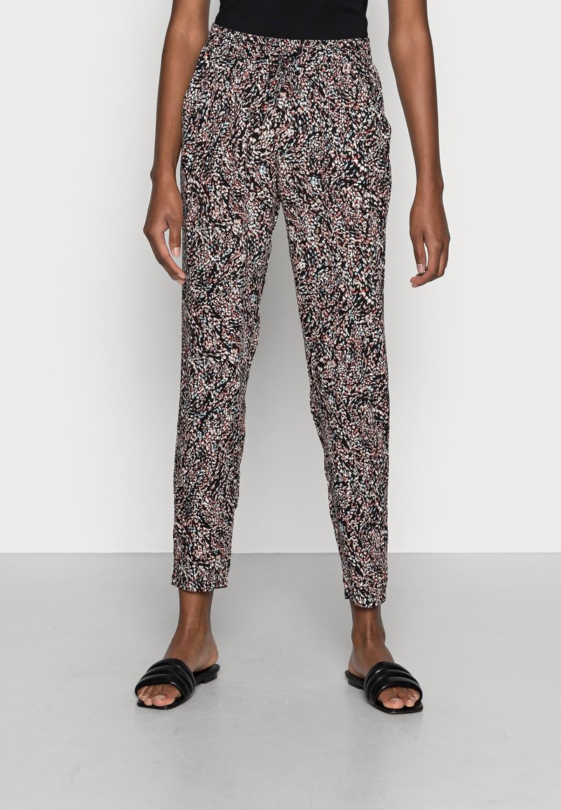 Femme portant un pantalon slim taille haute avec un imprimé abstrait noir, blanc et rose, associé à des sandales slides noires et un top noir.