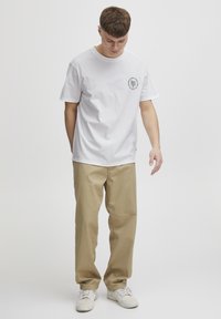 Hvid bomulds T-shirt med korte ærmer og rund hals; har et lille sort logo på brystet. Båret med beige bukser og hvide sneakers.