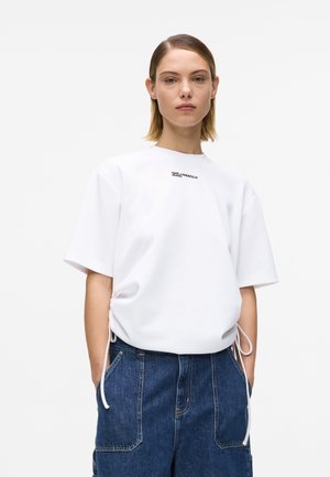 Fiatal nő rövid hajjal, fehér, oldalt megkötős rövid ujjú inget és kék farmert visel, egyszerű háttér előtt állva.