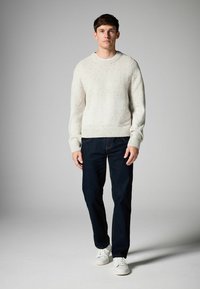 White knitted sweater with textured patterns, paired with dark blue jeans and white sneakers. Model stands against a gray background.