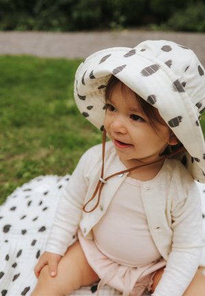 Bambino piccolo che indossa un cardigan bianco e un cappello da sole a pois, seduto su una coperta a pois abbinata all'aperto sull'erba.