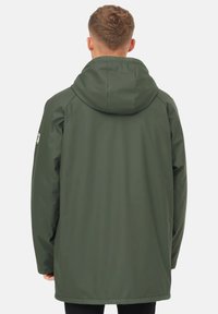 Osoba s krátkými, texturovanými vlasy stojí otočená zády k divákovi, na sobě má kapucňovou olivově zelenou bundu na jednobarevném pozadí.