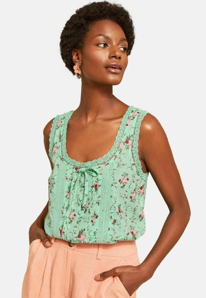 Woman with short curly hair wearing a light green floral sleeveless top and pale pink pants, looking to the side with hands in pockets.