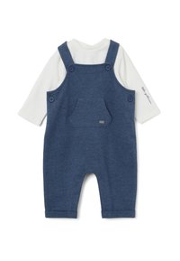 Denim-blaue Latzhose mit einem weißen Langarmshirt darunter. Sie verfügt über verstellbare Träger, zwei große Knöpfe und eine Fronttasche.