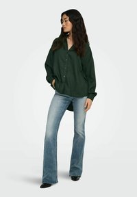 Green, oversized button-up shirt with a textured fabric, paired with light blue flared jeans and black pointed toe heels.