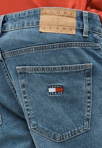 Blå denimjeans med tannfarget lærmerke med teksten "Tommy Jeans" på linningen og brodert logo på baklommen.