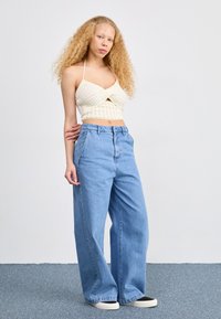 Top corto de punto amarillo claro con diseño cruzado, combinado con vaqueros de mezclilla azul de pierna ancha y cintura alta. Zapatillas blancas y negras.