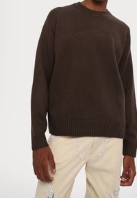 Brown knitted sweater with a crew neckline and ribbed cuffs, paired with beige pants featuring white leaf patterns and embellishments.
