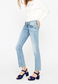 Ljusblå denimjeans med rak skärning, femfickorsdesign och subtil slitning, parat med marinblå klacksandaler och en krämfärgad topp.