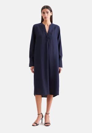 Femme aux cheveux raides et foncés portant une robe midi ample à manches longues bleu marine et des sandales noires à talons hauts, debout sur un fond blanc.