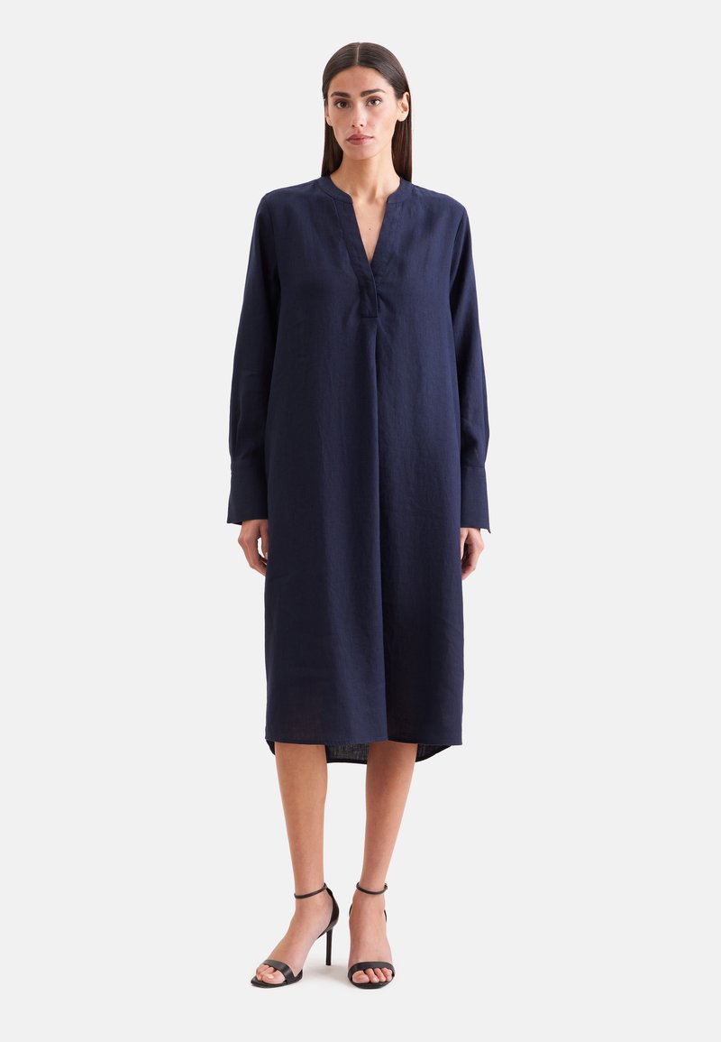 Femme aux cheveux raides et foncés portant une robe midi ample à manches longues bleu marine et des sandales noires à talons hauts, debout sur un fond blanc.