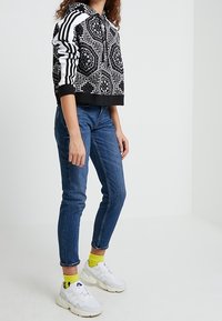 Černobílá vzorovaná crop mikina s kapucí, doplněná úzkými modrými džíny. Bílé tenisky s viditelnými žlutými ponožkami.