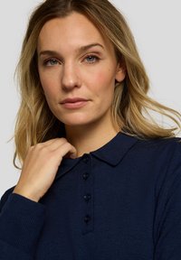 Dunkelblaues Poloshirt mit Kragen, fünfknöpfiger Knopfleiste und geripptem Stoff; das Modell berührt mit einer Hand den Kragen.