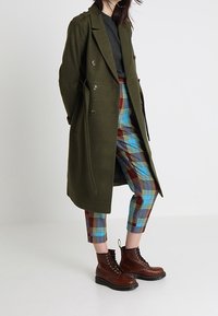 Olivgrüner, doppelt knöpfbarer Mantel mit Taillengürtel, getragen über einem grauen Oberteil. Blaue Karohose mit roten Akzenten und braunen Schnürstiefeln.