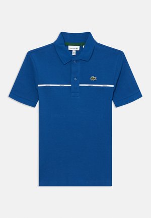Mėlyna polo marškinė, pagaminti iš tekstūruoto audinio, su dviem mygtukais priekyje, balta juostele su prekės ženklu ir žalia vidine apykakle. Žalias Lacoste logotipas.